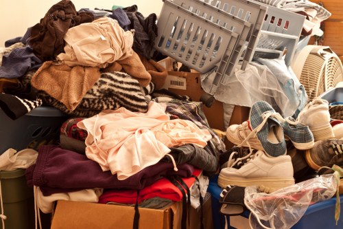 A large assortment of discarded household items and clothing piled together in a cluttered indoor space. The pile includes various textiles such as folded and unfolded garments, blankets, and towels in different colours and fabrics. Among the clothing are shoes, some in pairs and others scattered, with a mix of casual footwear and slippers, situated towards the front right of the image. There are also plastic storage crates, some stacked, containing additional items, and a cardboard box partially visible beneath the pile. In the background, the edge of a piece of furniture or a container can be seen, with the environment suggesting an area used temporarily for storing rubbish or unwanted belongings. The lighting appears natural, illuminating the jumble of objects, highlighting their textures and colours. This scene exemplifies clutter that a rubbish removal service such as House Clearance Oxted might manage, especially in a residential setting within or near Oxted postcode areas.