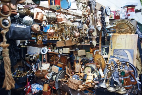 A densely packed outdoor display of various discarded household items, including copper and brass cookware, metal lamps, and ornamental objects, arranged on racks and tables. The assortment includes different textures and finishes, such as polished, tarnished, and rusted surfaces, with objects overlapping and stacked closely together. Some items, like a vintage clock and a brass tray, are positioned prominently at the front, while others, such as small decorative items and utensils, fill the background and gaps. The setting appears to be a flea market or a second-hand shop, with a mix of antique and used items showing signs of wear. The lighting suggests natural daylight, subtly highlighting the metallic and wooden textures of the objects. This scene reflects a collection of miscellaneous rubbish suitable for professional waste removal, akin to services offered in Oxted and nearby areas, with a focus on metal and household clutter.
