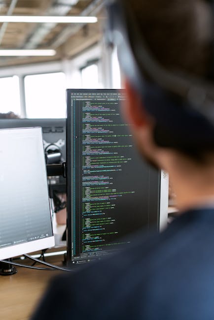 A person with short, dark hair and wearing a dark top is working in an office environment, focused on a large computer monitor displaying lines of multicoloured code on a black background. The workspa