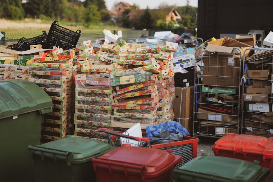 A large pile of mixed waste is situated on a gravel surface outdoors, consisting of black plastic rubbish bags, cardboard, and loose debris. Prominently, a worn, beige car tire lies on its side in the